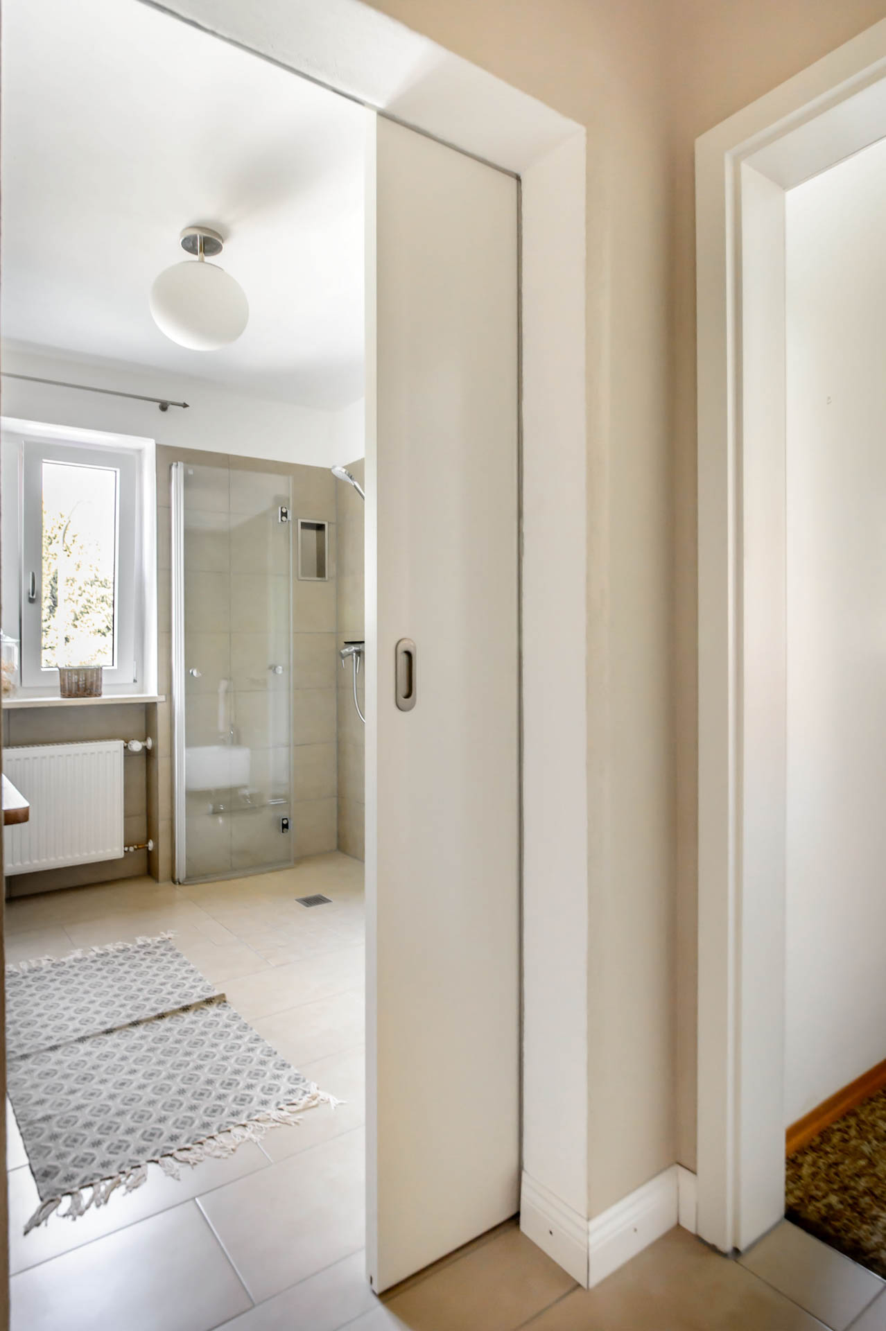 Blick aus dem Flur durch die halb offene Schiebetür in das helle Badezimmer mit Fenstern und Glas-Duschkabine.
