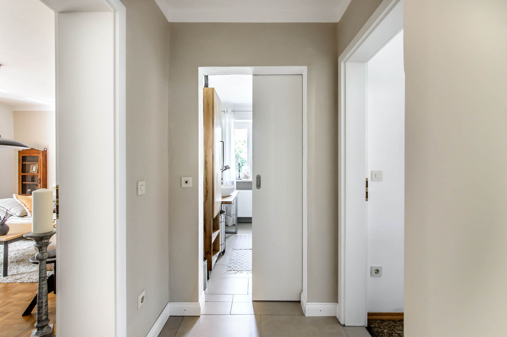 Flur mit beiger Wandfarbe und offener weißer Schiebetür, die den Blick in ein helles Zimmer dahinter mit Fenster freigibt.