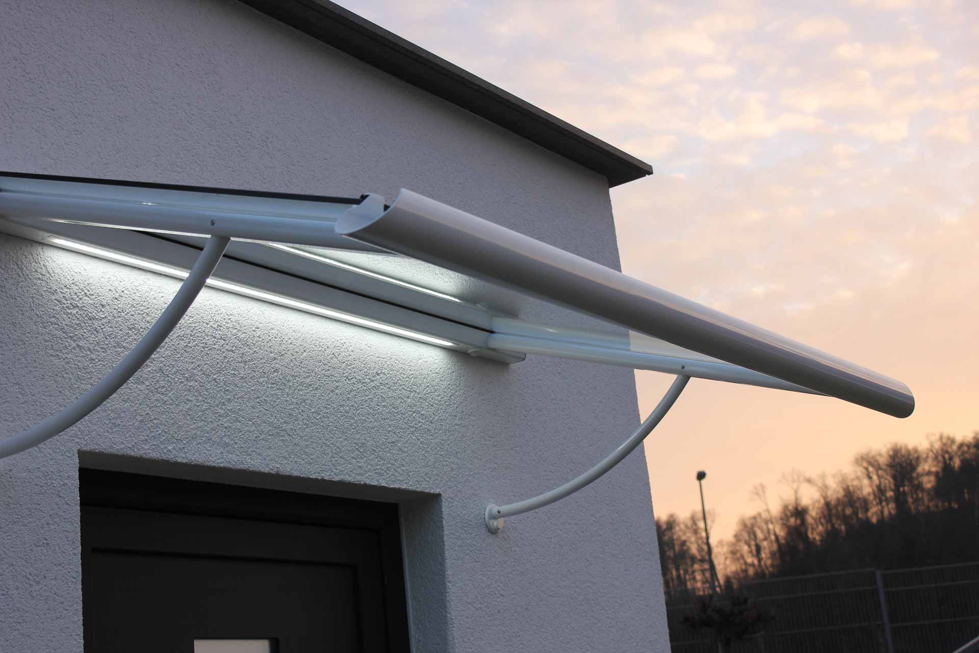 Elegantes, weißes Vordach mit gebogenen Haltearmen und integrierter LED-Beleuchtung vor Abendhimmel.