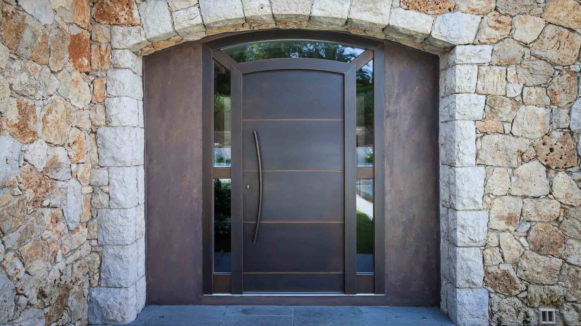 Geschwungene, dunkelbraune Eingangstür mit Rundbogen-Glasausschnitt in rustikalem, sandsteinfarbenen Mauerwerk.