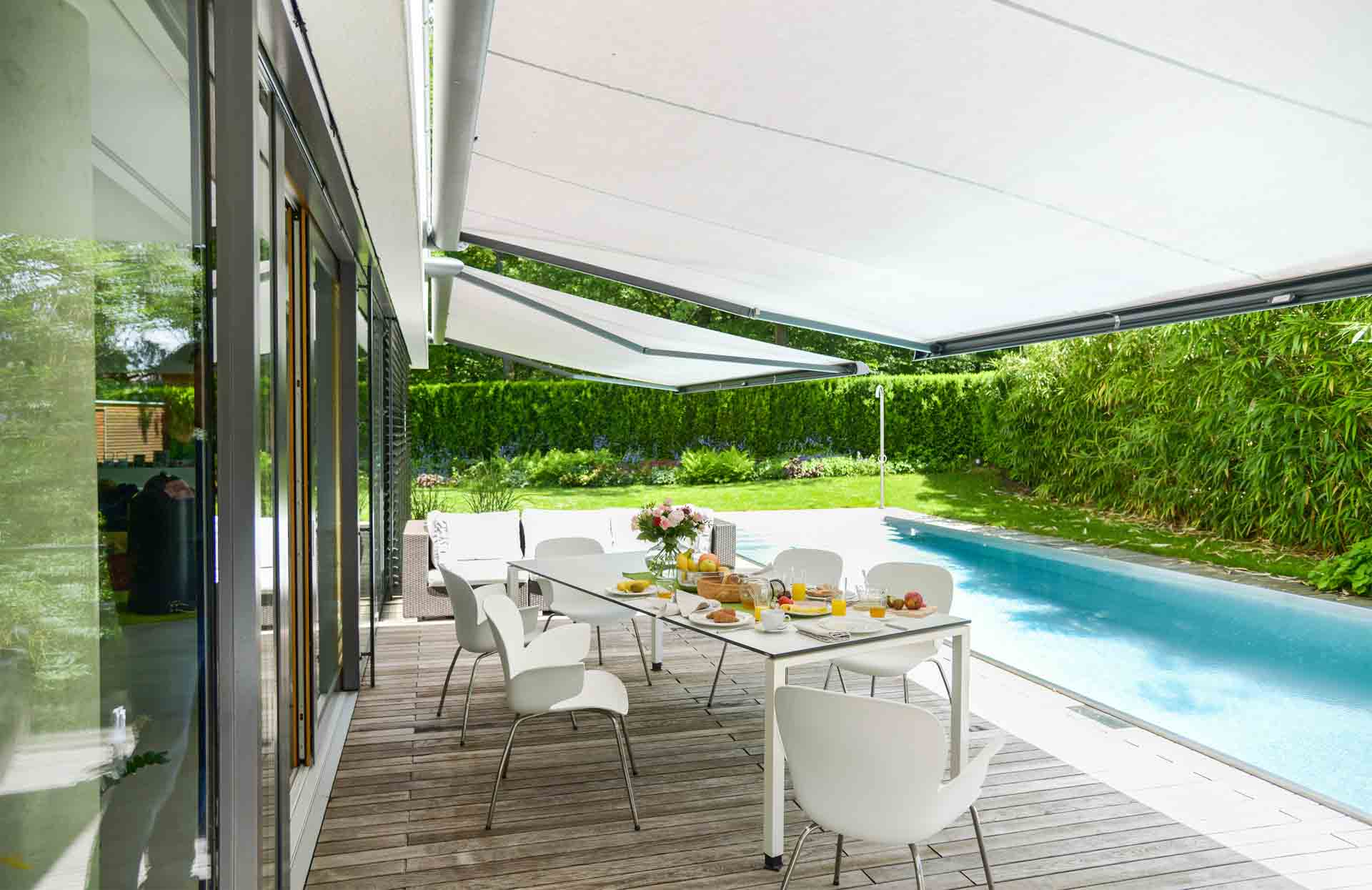 Große, weiße Markise spendet Schatten über einer Holzterrasse mit Esstisch und Pool im Garten.