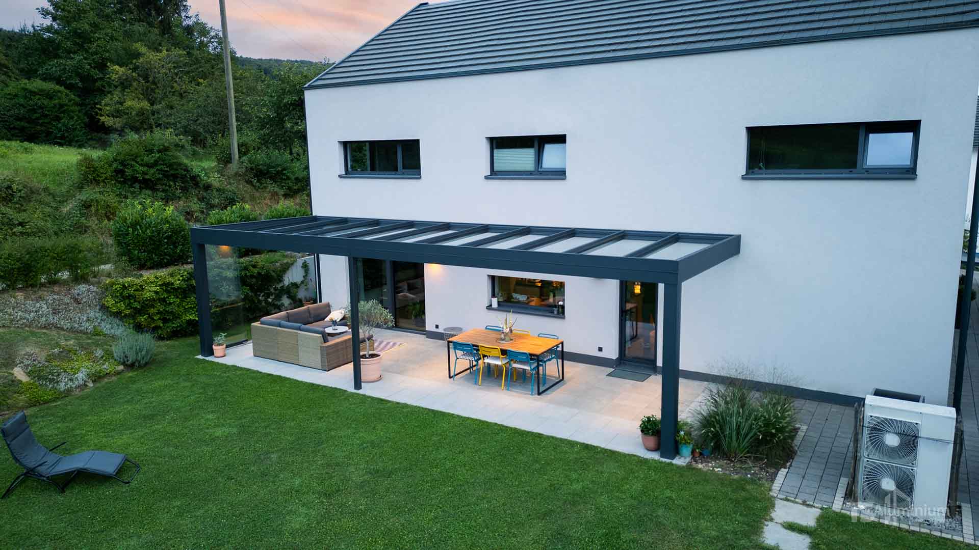 Dunkles Aluminium Terrassendach mit Glasdach über der Terrasse eines modernen Einfamilienhauses im Grünen.