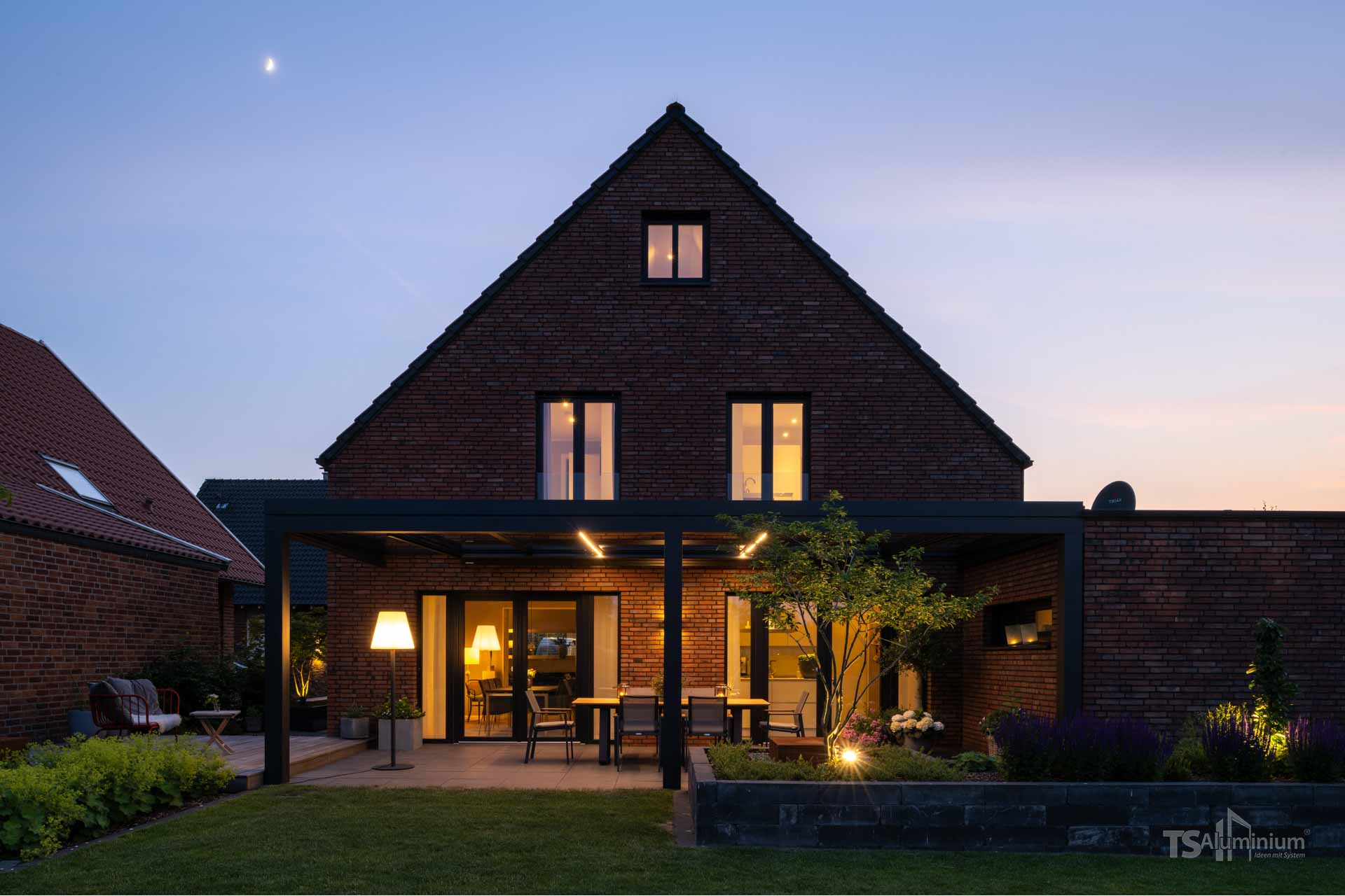 Schwarzes Terrassendach mit integrierter Beleuchtung erweitert die Terrasse eines klassischen Backsteinhauses am Abend.